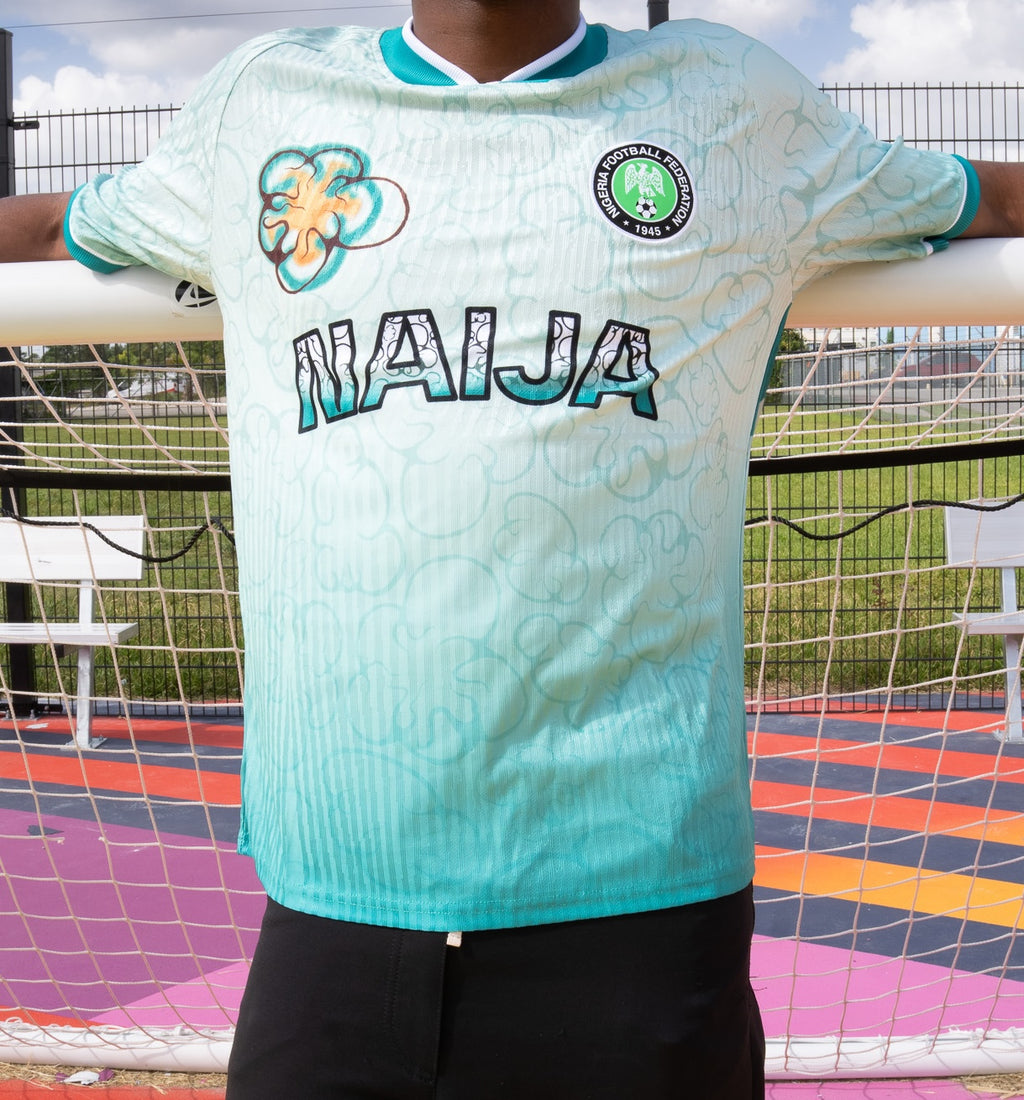 A person wearing a light aqua soccer jersey with 'NAIJA' across the chest, a floral crest on the right, and a circular club crest on the left, photographed outdoors in front of a soccer goal.